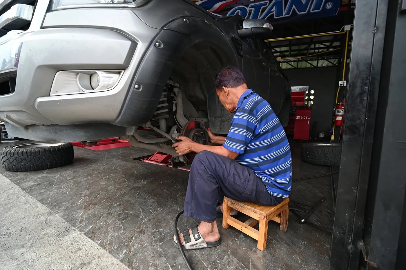 Mechaniker auf einem Holzhocker arbeitet an der Vorderachse eines angehobenen Pickups